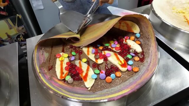 A close-up shot capturing a colorful rainbow crepe being expertly folded using a metal spatula on a hot commercial griddle.