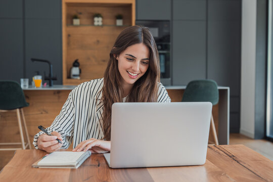 Woman studies online with a laptop and notebook at her kitchen table, writing lesson notes with a pen. Smiling student supports online learning, education, homework, research and study from home.