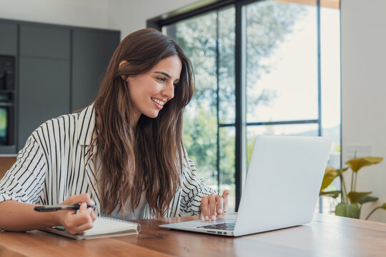 Woman searches for a job on her laptop while holding a pen over a notebook at home. Smiling job seeker explores career opportunities, application tasks, employment and interview preparation online.