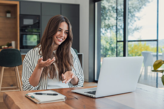 Woman has an online doctor consultation on her laptop at the kitchen table, smiling as she describes her concern. Her gesture supports telehealth, virtual healthcare, remote care and digital health.