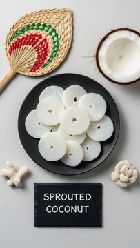 Minimalist flat lay of traditional Marshallese Iu sprouted coconut slices on a dark ceramic plate with a tiny chalkboard, a hand-woven fan, half a coconut shell, and sun-bleached coral.