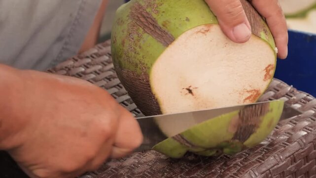 close-up shot capturing a person's hands skillfully slicing into a vibrant fresh green coconut using a sharp cleaver knife.
