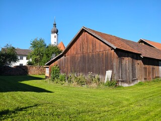 Rustic wooden barn beside village church tower above green lawn under blue sky © Waldemar