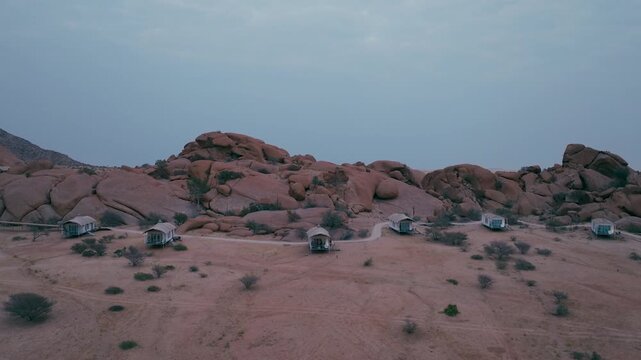 An aerial drone shot revealing a row of sustainable glamping tents nestled against rocky mountains in the Namibian desert during twilight. Perfect for travel and ecotourism content.