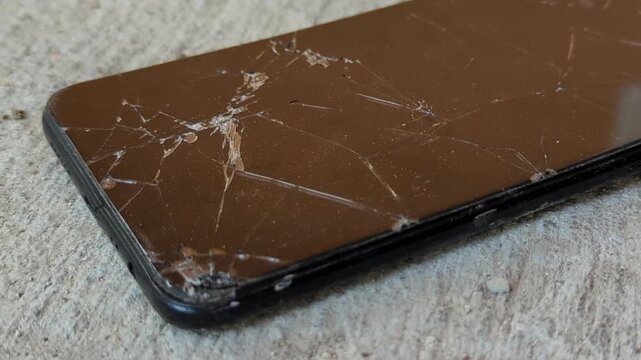 Heavily damaged smartphone with a severely cracked and shattered glass screen lying face up on rough concrete ground.