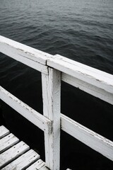 Fototapeta premium White wooden fence and pier railing over dark ocean water. Old weathered wood structure stands near sea. Close up view of maritime construction on coastal boardwalk during summer.