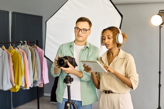 Photographers team review camera tablet in studio. Discussing shots near lighting and a clothes rack, they plan fashion photoshoot project and equipment. Clear creative collaboration concept.