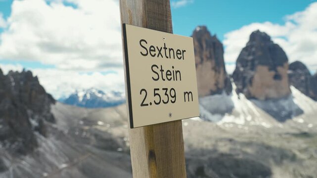Wooden summit sign reading Sextner Stein 2539 m on a post with the blurred Tre Cime di Lavaredo north faces and rocky slopes in the background in the Dolomites Italy