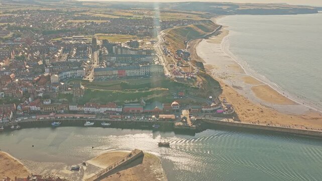 Whitby West Cliff overlooks Whitby Sands and the River Esk estuary, with the harbour, beach and dense seaside town extending along the North Sea coast, North Yorkshire, England, drone forward flyover
