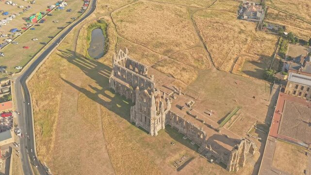 Whitby Abbey Gothic ruins dominate the East Cliff above Abbey Plain, with the medieval church, surrounding grounds and Whitby town beyond in North Yorkshire, England, drone forward flyover, descent