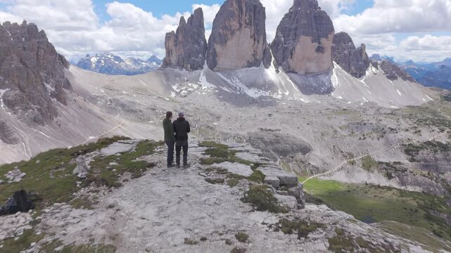 Wide aerial drone view of two hikers standing together on an open rocky plateau facing the Tre Cime di Lavaredo north faces with snow patches and mountain ranges around them in Italy