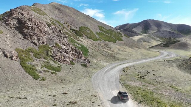drone perspective following an automobile navigating a gravel road flanked by towering rocky slopes and arid landscapes during a road trip in Yol Canyon