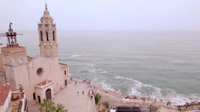 4K drone aerial view of Sitges, Spain, featuring the iconic Sant Bartomeu i Santa Tecla church overlooking the Mediterranean coast, beaches and historic seaside town.