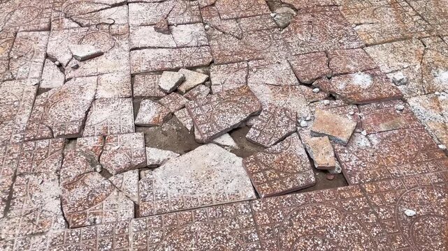 Broken and heavily cracked ceramic tiles on a damaged sidewalk pavement. A close-up view of shattered floor tiles showing gaps, loose debris, and exposed soil beneath the surface outdoors. 