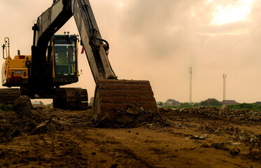 Excavator bucket coated with soil and resting on uneven ground at a housing estate construction site, attached to a hydraulic arm, with crawler tracks, a yellow cab and distant houses beneath clouds. © Fahroni