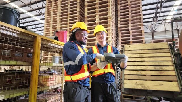 Two protected factory workers discuss pallet production and inspect equipment inside a busy industrial warehouse filled with stacked wooden pallets.