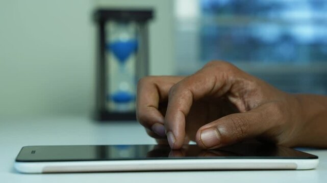 Hand using tablet beside blue sand timer, Close-up of a hand using a tablet on a desk with a blue sand timer in the background, showing time management, technology, and work focus.