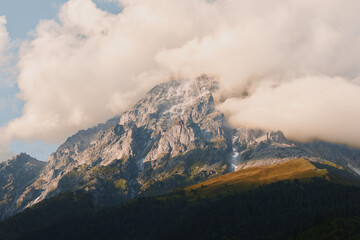 Naklejka premium Majestic mountain peak rising above a pine forest, with wispy clouds, rugged rock, and expansive alpine landscape highlighting nature, scale, and solitude and calm mood.