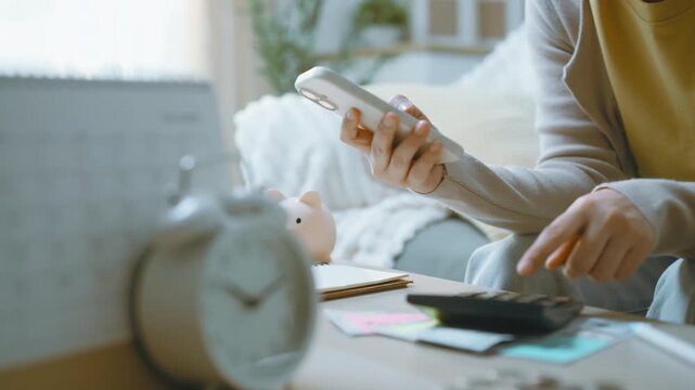 Calculator use beside a smartphone illustrates finance and budget planning at home, with an alarm clock and piggy bank nearby as hands combine button presses with a finance review at the table.