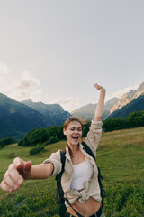 Naklejka premium A joyful woman hikes through a green valley with distant mountains and a backpack, capturing a candid outdoors lifestyle moment of energy, freedom, and exploration.