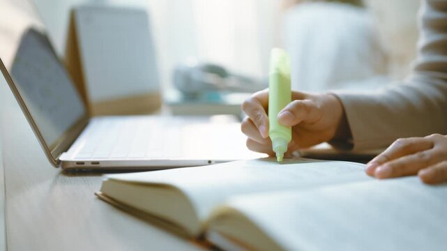 Highlighting text with a green highlighter shows reading and study beside an open book, with one hand holding the page as highlighting emphasizes a passage near a laptop in a domestic workspace.