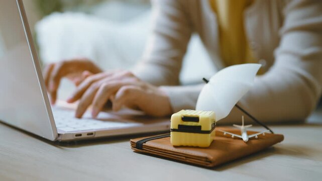 Travel planning is represented by hands typing on a laptop behind a miniature suitcase and airplane, with the trip accessories on a notebook as travel preparation becomes a computer activity.