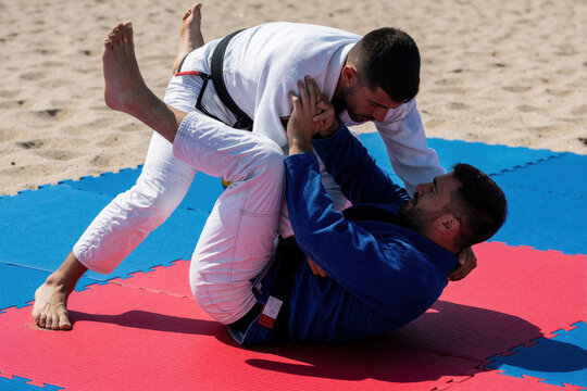 Two men practice jiu jitsu on beach mats. Athletes in blue, white gi fight during outdoor training session. Young men engage in grappling martial arts combat on sand under summer sun. Strong men