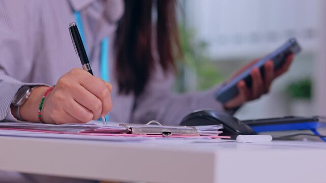 Woman Writing Business Paperwork at Desk While Holding Smartphone and Pen, Recording Information on Document in Bright Modern Office