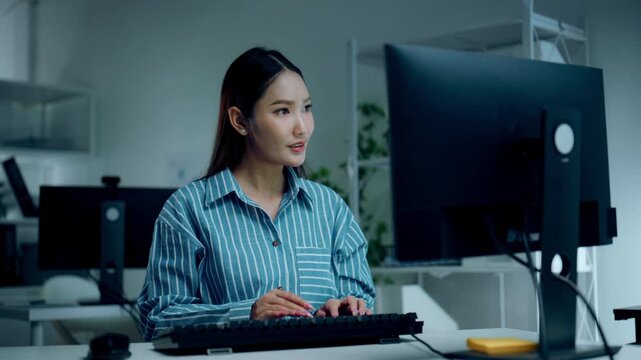 Typing at an office computer, a woman in a blue striped shirt is working with her gaze toward the keyboard, showing a slight smile as the woman carries out an seated task in the dim workspace.