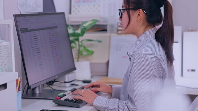 Woman Office Worker Typing on Computer Keyboard at Desk, Reviewing Spreadsheet Data on Monitor in Bright Workspace with Plant and Office Supplies