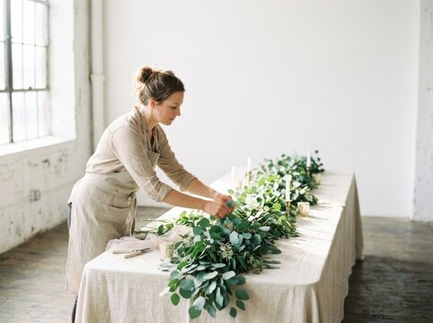 A woman arranges greenery on a long table in a minimalist, bright room with large windows. She is wearing a beige apron and has her hair tied up. The table is covered with a white tablecloth 