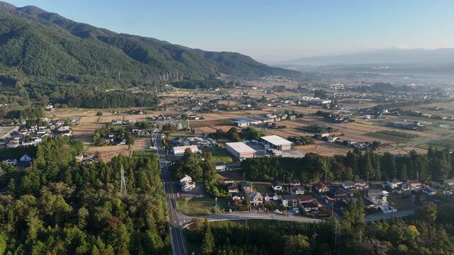 An aerial view shows low buildings, rectangular fields and a cluster of large light colored roofs beneath forested mountains.