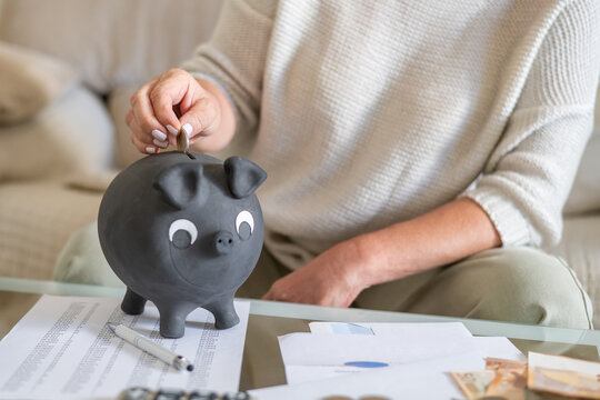 Woman's hand depositing a coin into a black piggy bank, managing financial savings, household bills, and budgeting to control expenses and combat rising inflation