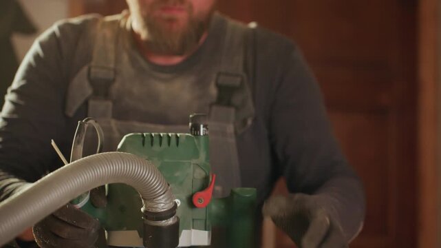 Close up shows upper body and hands of bearded worker in overalls using power tools for wood machining. scene focuses on manual operation protective gear ear muffs and industrial lighting