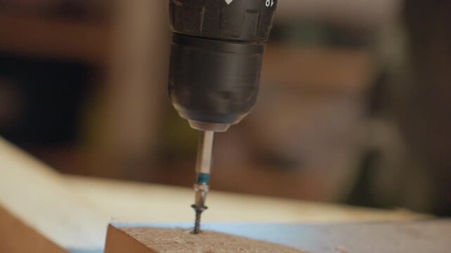 Close up shows drill driving screw through wooden board with steady motion focusing on precision of power tool and material interaction. Drill bit engages wood surface creating clean hole