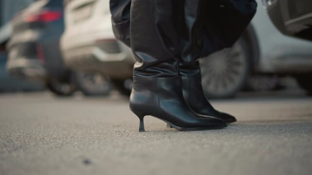 Close up shows pair of feet wearing sleek black leather boots standing and slightly shifting position over urban street scene. focus remains on texture shape and movement of footwear