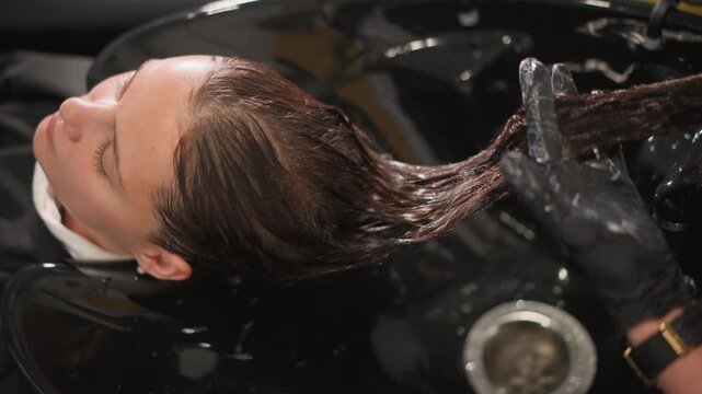 Client lies back in black basin as gloved hand applies and works through dark wet hair during coloring treatment stylist care perm process trimming scissors