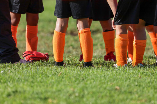 A low-angle, close-up shot of the lower legs and feet of a youth soccer team standing together on a lush green grass field.