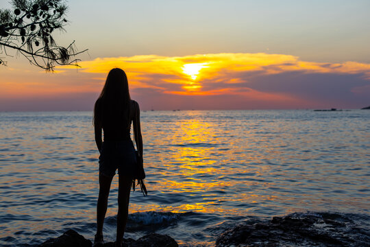 Silhouette of a young woman standing on a rocky seashore watching a beautiful golden sunset, peaceful summer evening with colorful sky and sunlight reflecting on calm sea.