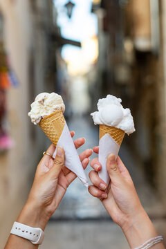 Close up of two hands holding delicious scoops of creamy gelato in crispy waffle cones outdoors, sweet summer ice cream treats against a blurred narrow European street.