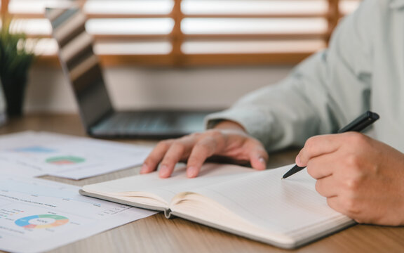 Business person writing notes in a notebook on a wooden desk with financial charts and a laptop in a modern office. Professional planning, data analysis, and corporate strategy concept.