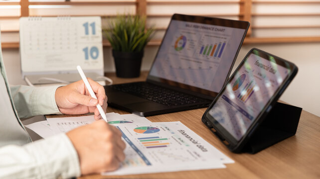 Professional person writing notes on financial chart documents with a digital tablet and laptop in a modern office. Data analysis, corporate planning, and business growth concept