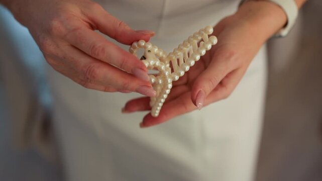 Close up view of hands holding and examining elegant white pearl hair clip. subject manipulates beaded object highlighting its texture detail while wearing light colored outfit