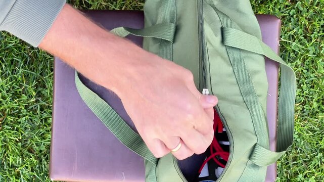 Top view of hands placing gear, smartphone, gorilla tripod, and cables into a green canvas bag on a bench outdoors on grass. Content creator gear preparation.