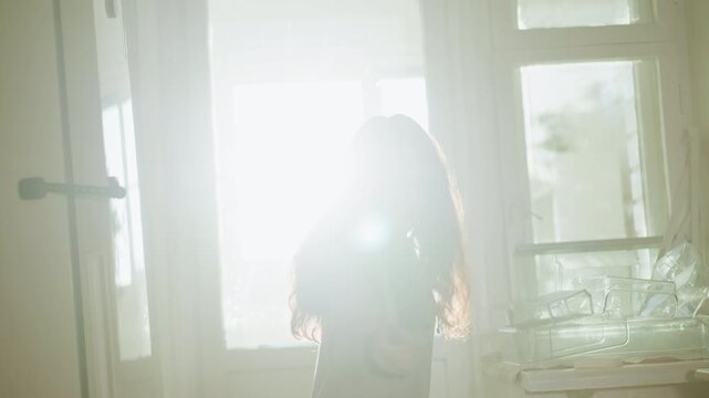 Woman stands silhouetted by sunlight moving slightly while interacting with two long items. Bright natural lighting fills room where clear glass containers are visible on surface