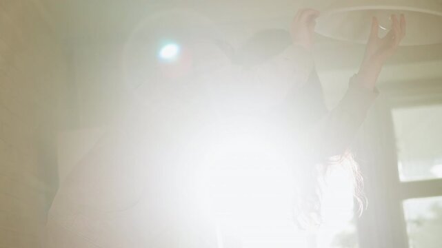 Woman reaches upward adjusting bright white circular lighting fixture on ceiling. Sunlight streams through large windows behind her illuminating soft indoor details