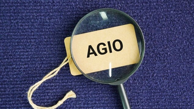 A magnifying glass and a paper tag with the word Agio is a financial term that refers to the extra cost, premium, or percentage paid for exchanging one currency for another, or the difference between 