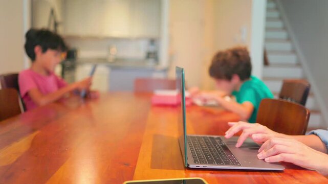 Mother working from home on a laptop with her children studying and using a smartphone at the kitchen table, highlighting the challenges of modern family life and remote work