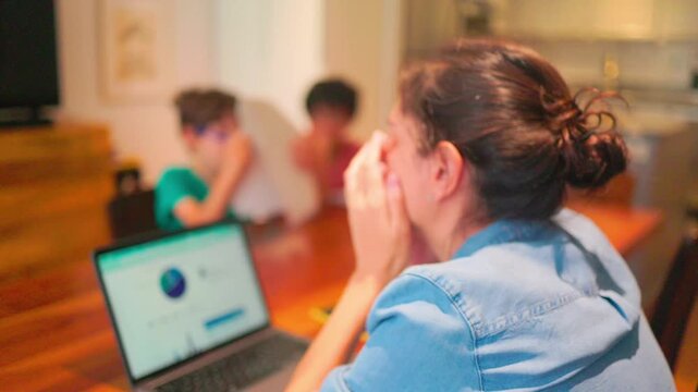 Stressed mother working from home experiencing burnout while her children are playing in the background, showing the challenges of remote work and family life
