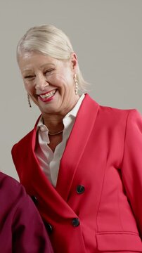 Vertical video: Smiling older woman in red blazer leaning laughing as pal entering frame in studio